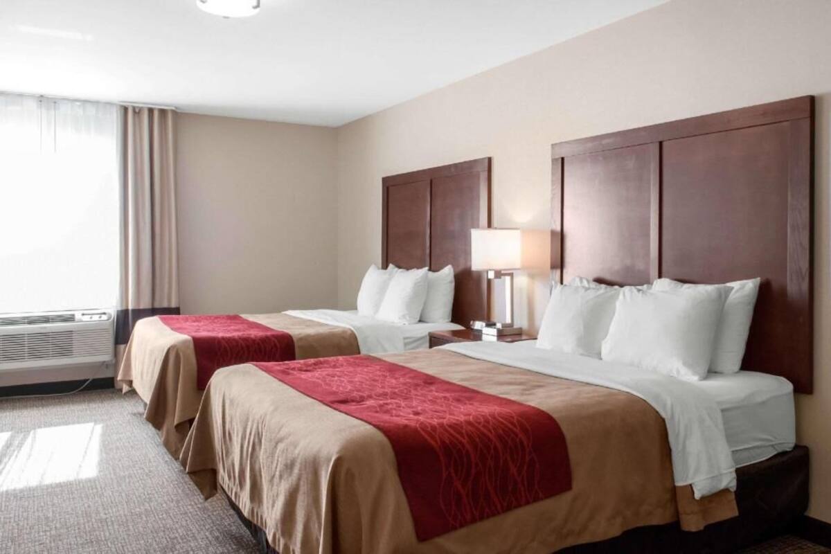A hotel room with two beds, a window with curtains, a bedside lamp, and a wall-mounted air conditioning unit.