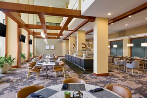 Un comedor de hotel amplio con mesas y sillas largas, una barra central y ventanas grandes.