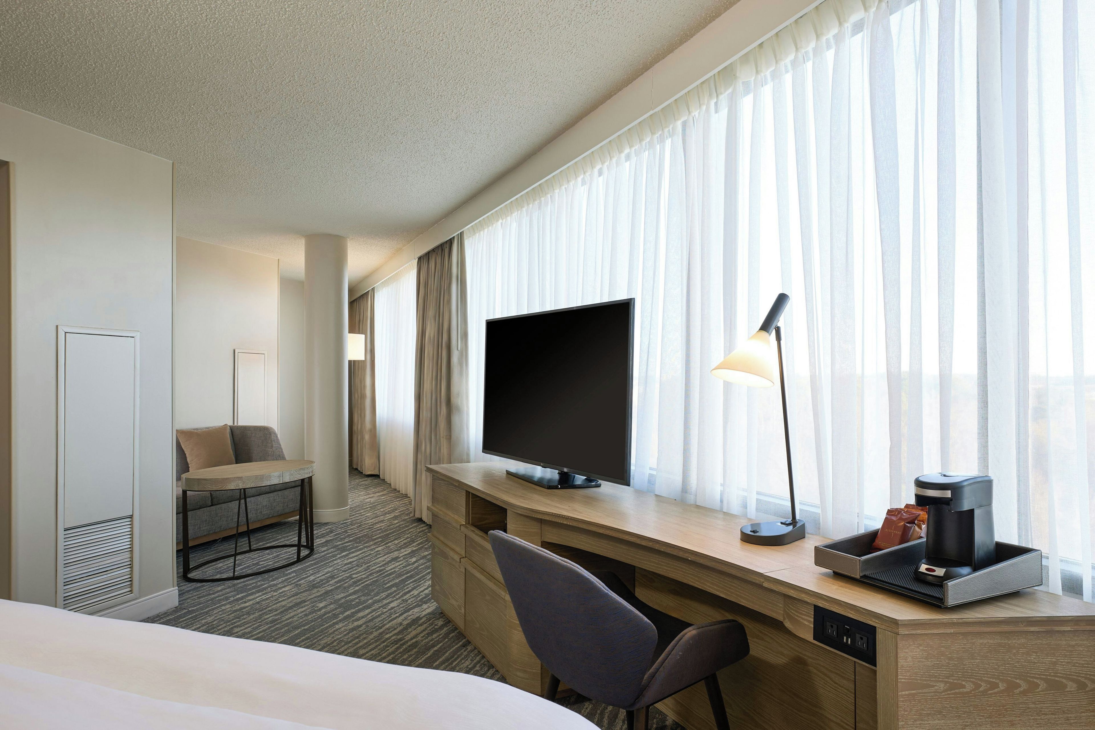 A hotel room with a bed, a desk with a flat-screen TV, a chair, a lamp, and a coffee maker.