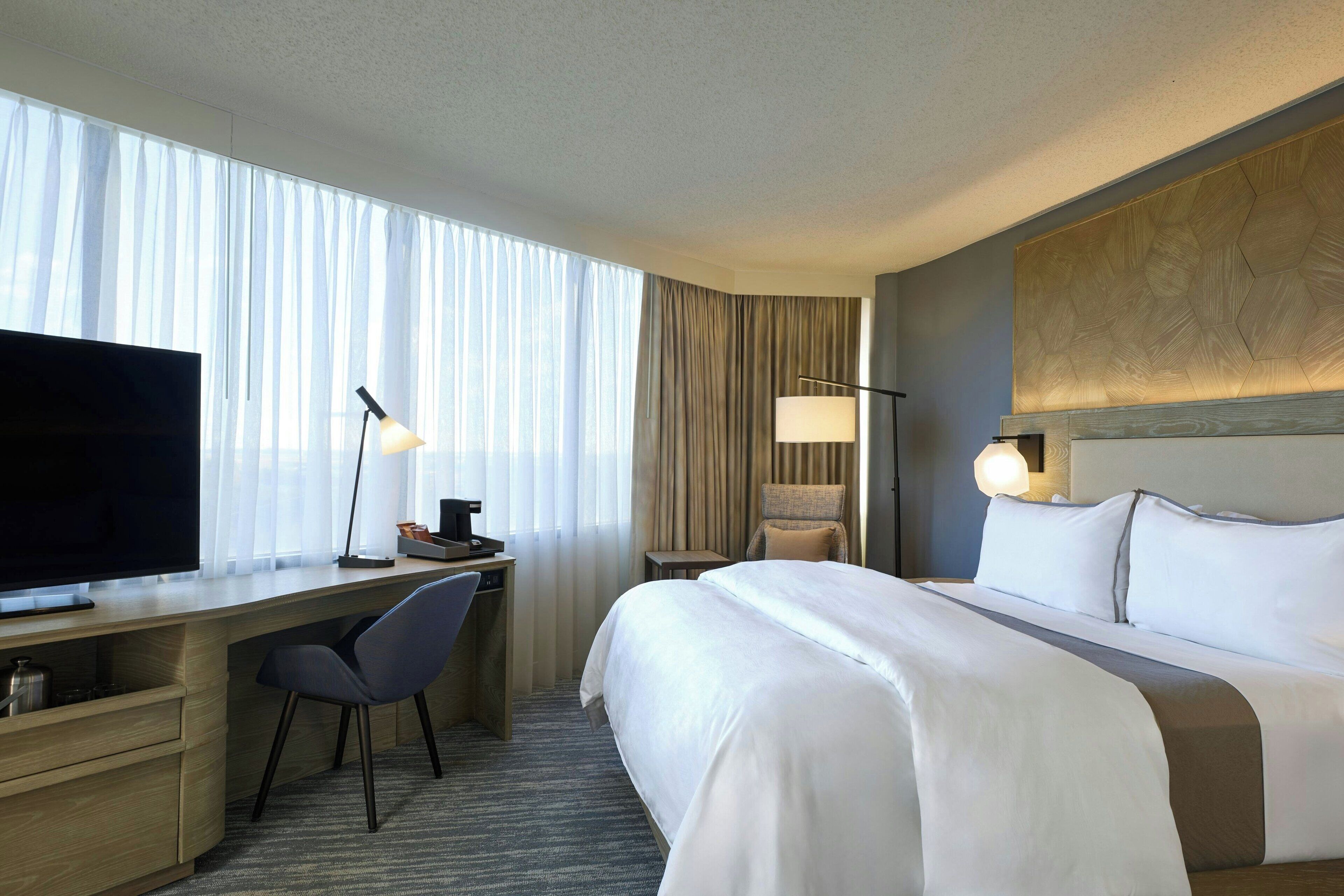A hotel room with a large bed, a desk with a TV, a chair, and a lamp.