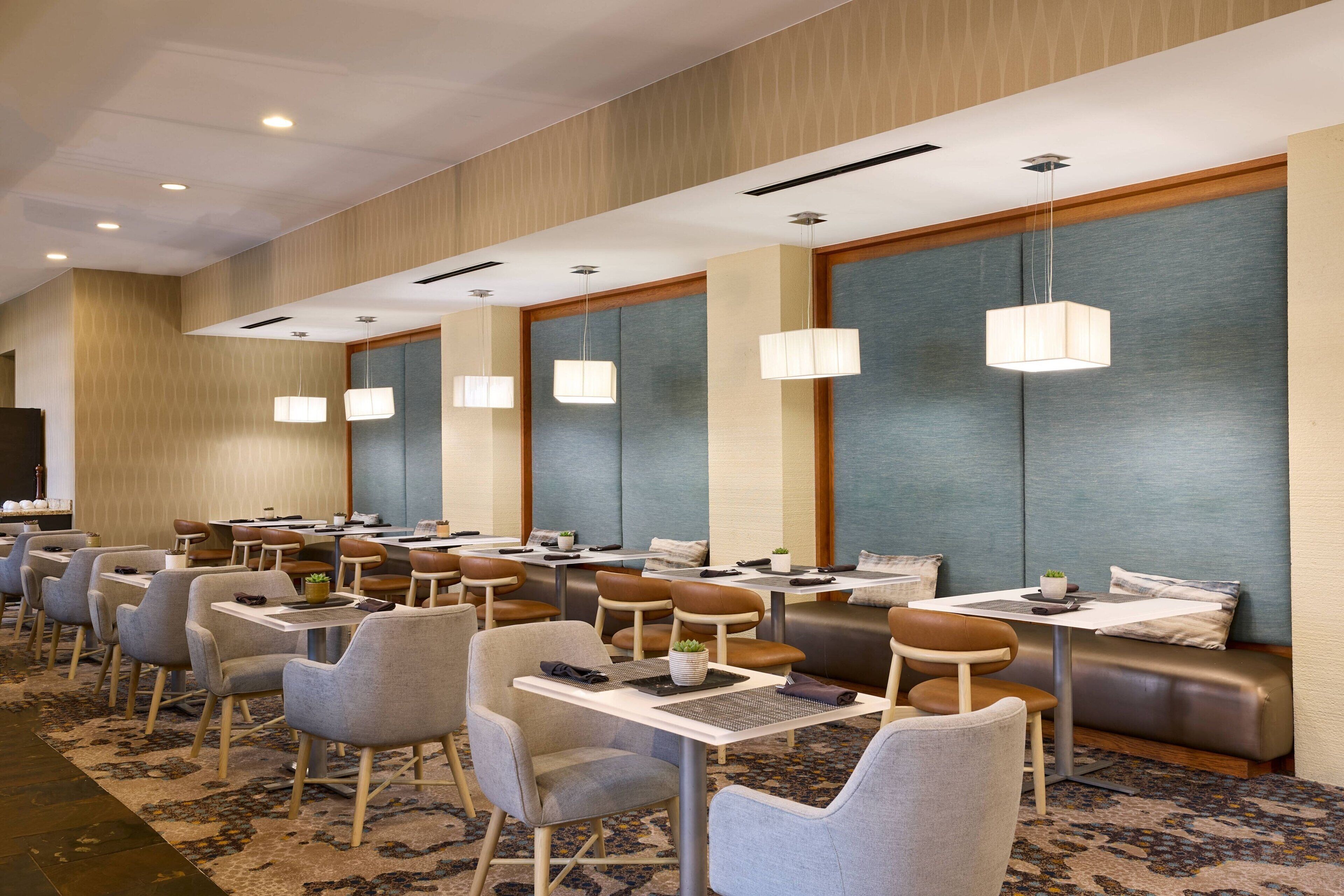 Comedor de hotel con mesas y sillas, iluminación empotrada y alfombra estampada.