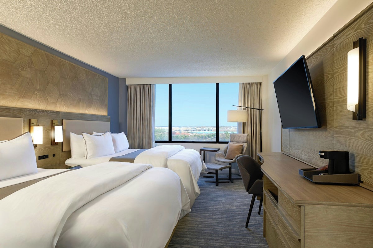 A hotel room with two beds, a desk, a chair, a TV, and a large window offering a city view.