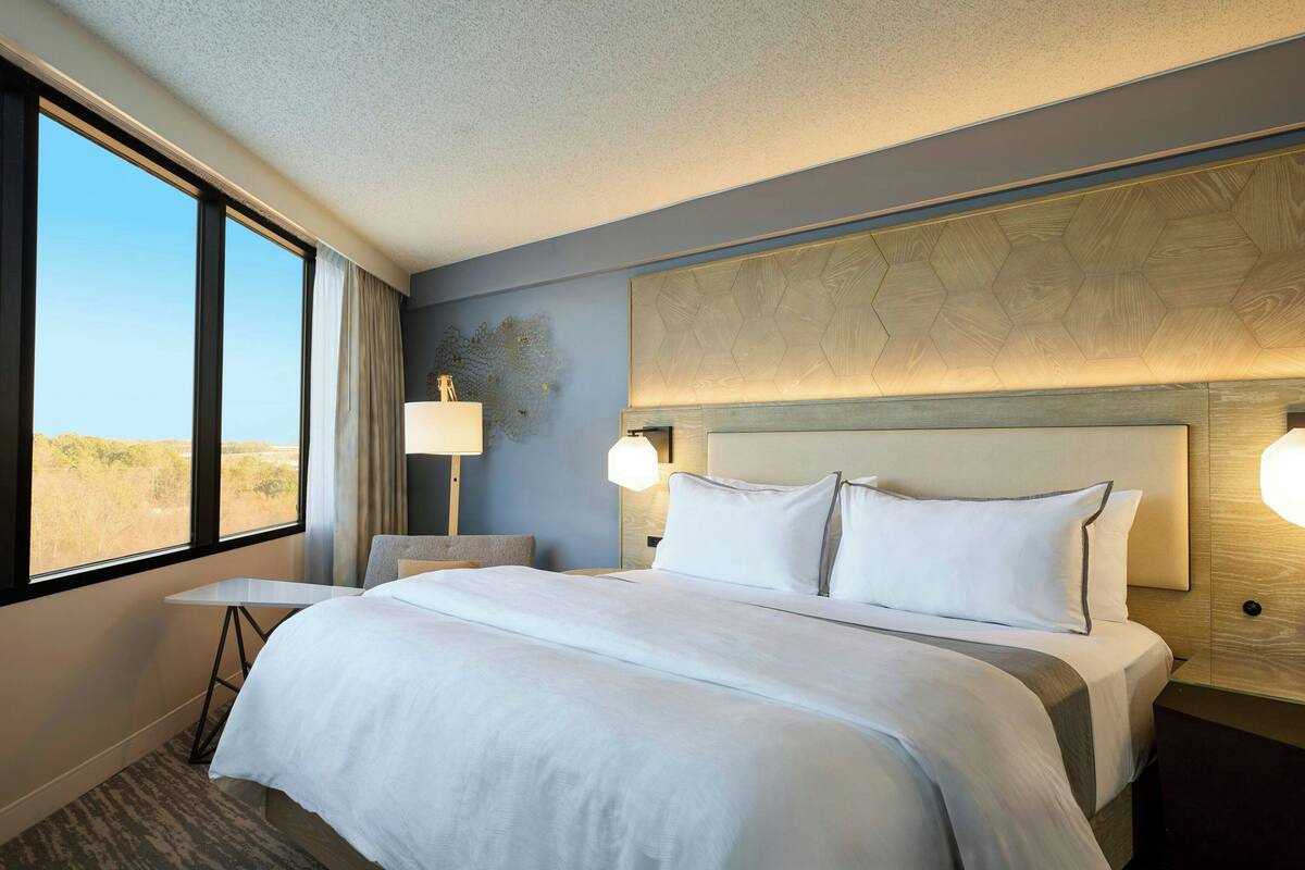 A hotel room with a large bed, a small table, a lamp, and a view of the desert through the window.
