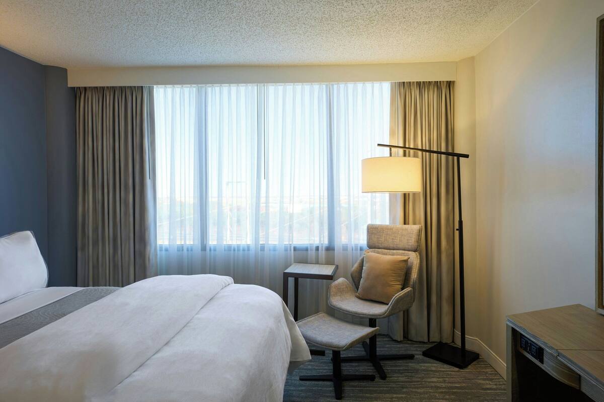A hotel room with a large bed, a chair, a desk, and a floor lamp.