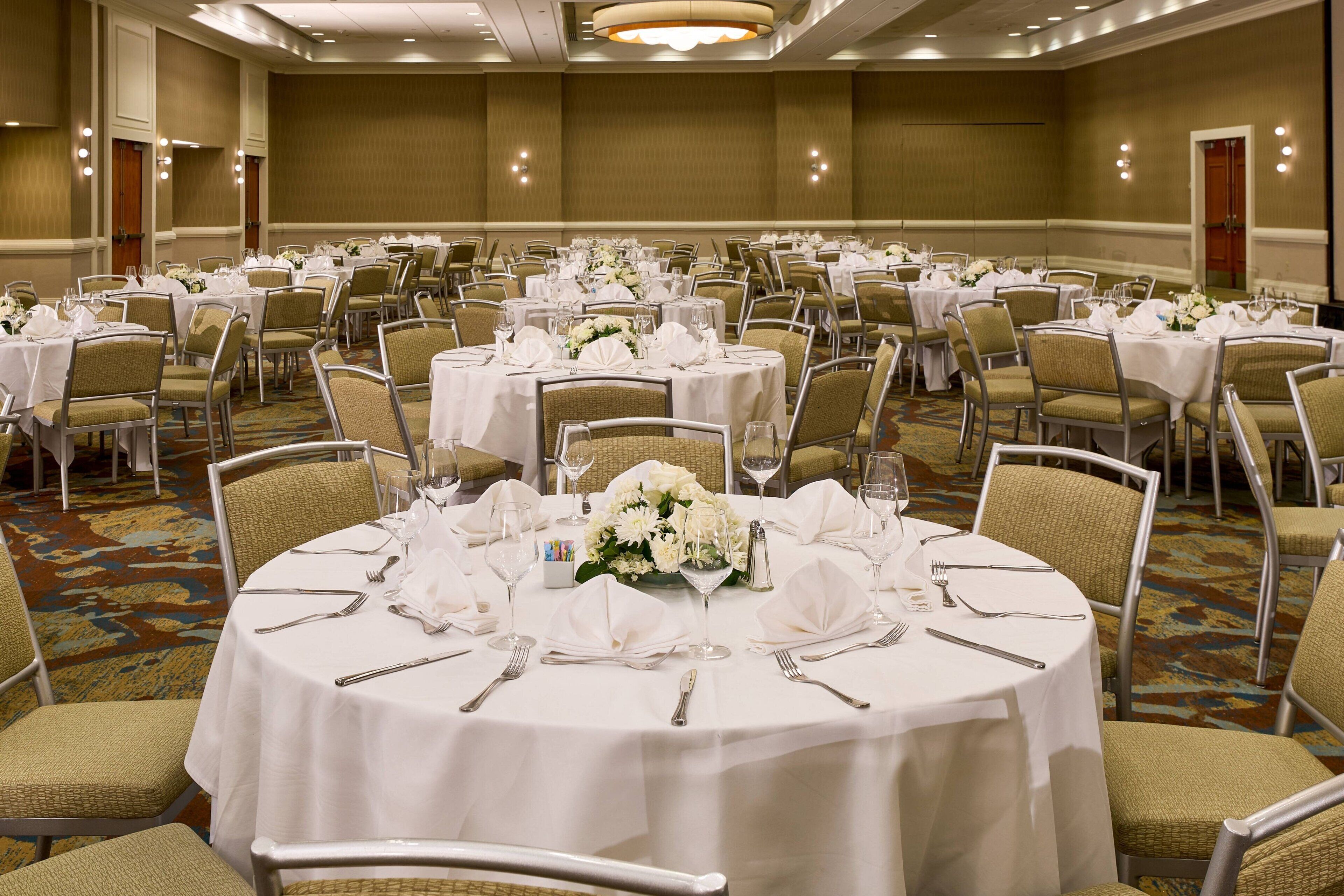Une salle de banquet avec des tables rondes dressées pour le repas, chacune étant recouverte d’une nappe blanche et agrémentée de chaises et de décorations de table.