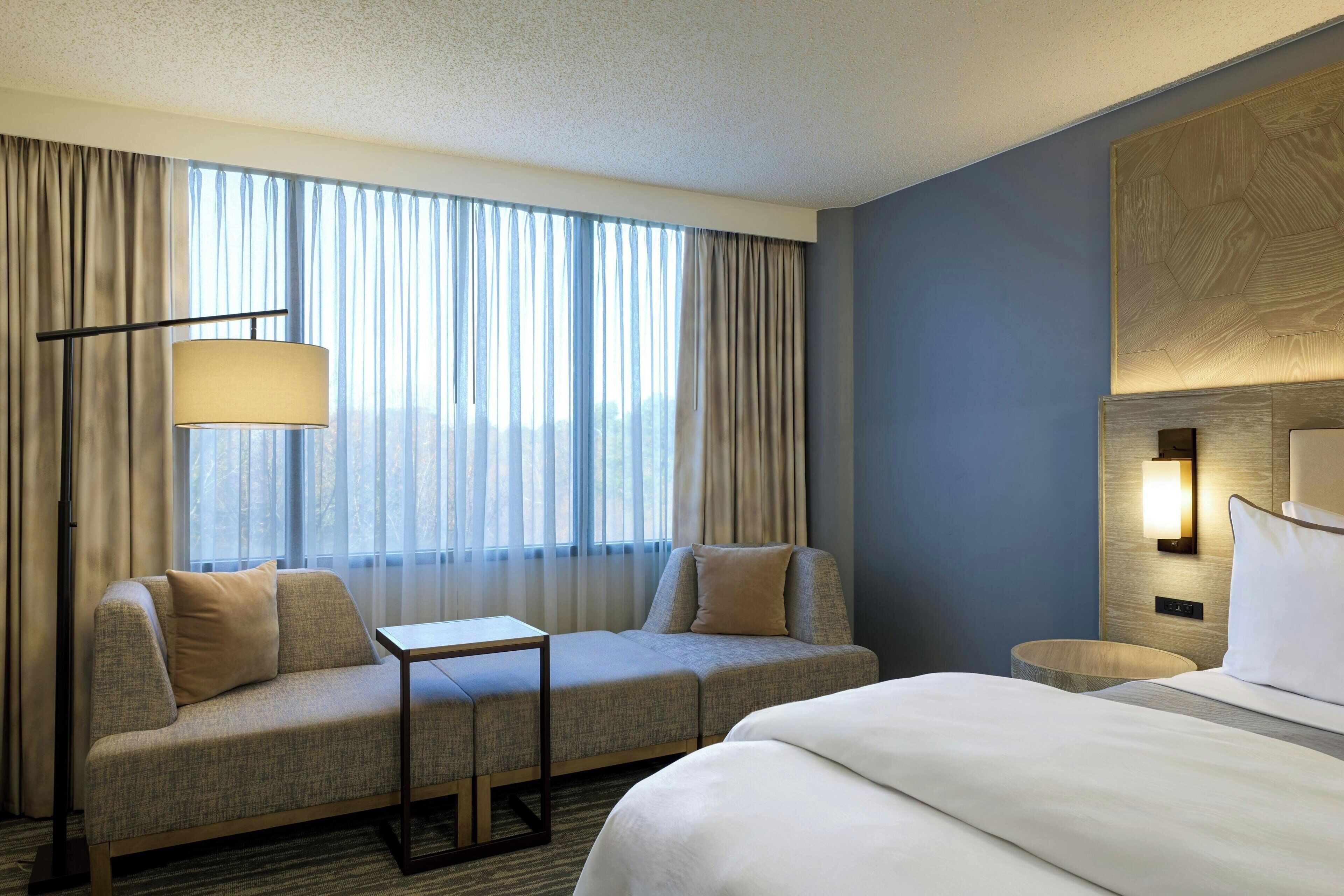A hotel room with a sofa, a small table, a bed, a lamp, and a view of the outdoors through large windows.