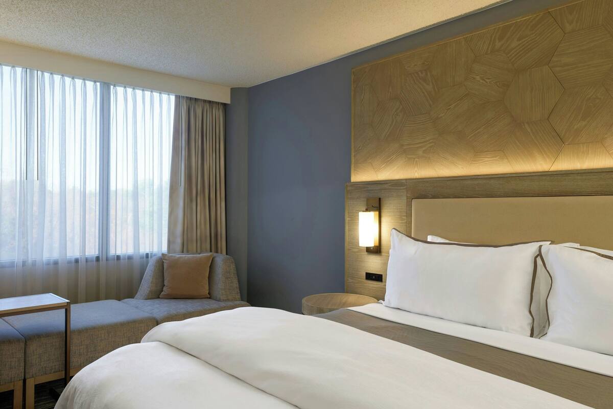 A hotel room with a large bed, a seating area with a sofa and armchair, a bedside table, and a wall-mounted light fixture.