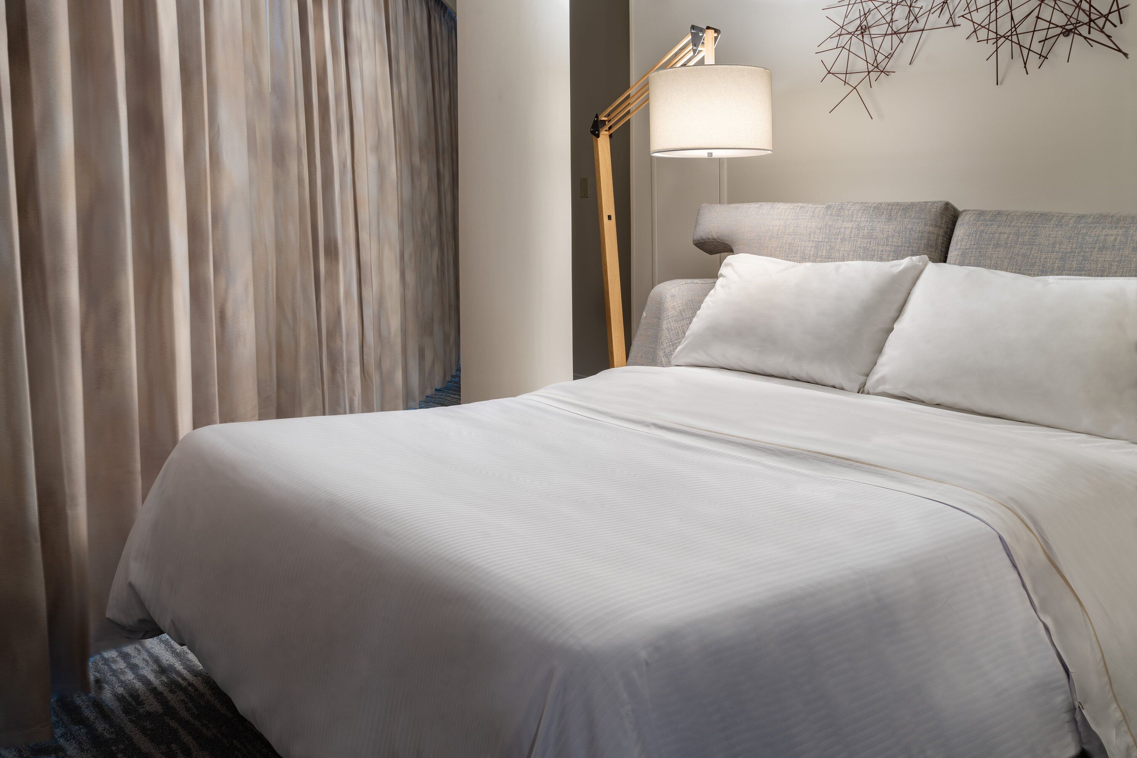 A neatly made bed with white linens, a floor lamp, and a wall art piece.