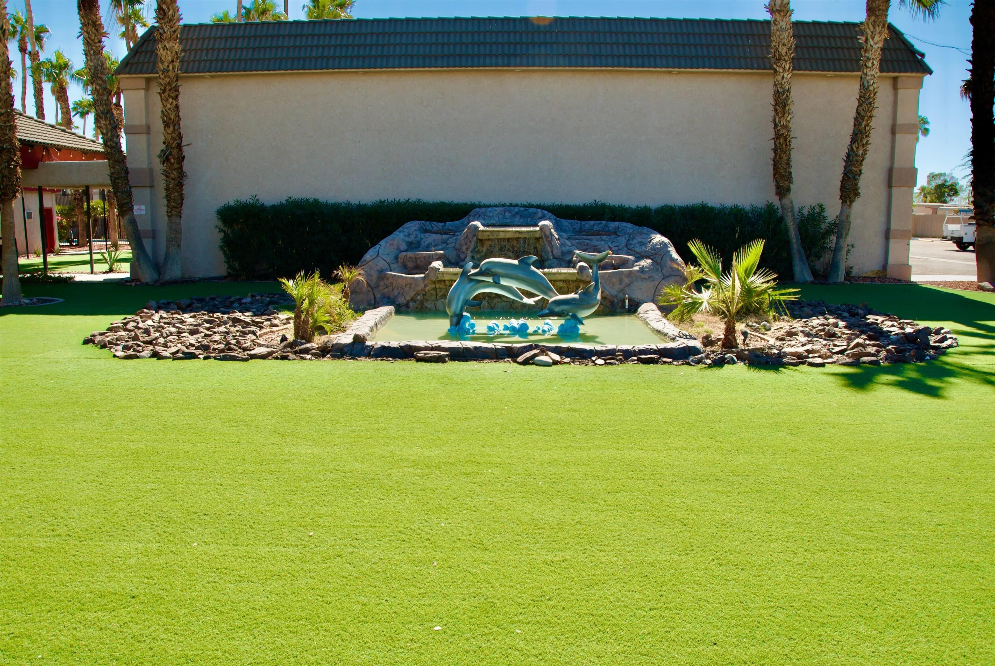 A building with a fountain sculpture in the center, surrounded by a well-maintained lawn and palm trees.
