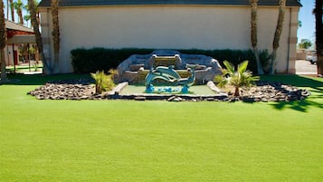 A building with a fountain sculpture in the center, surrounded by a well-maintained lawn and palm trees.