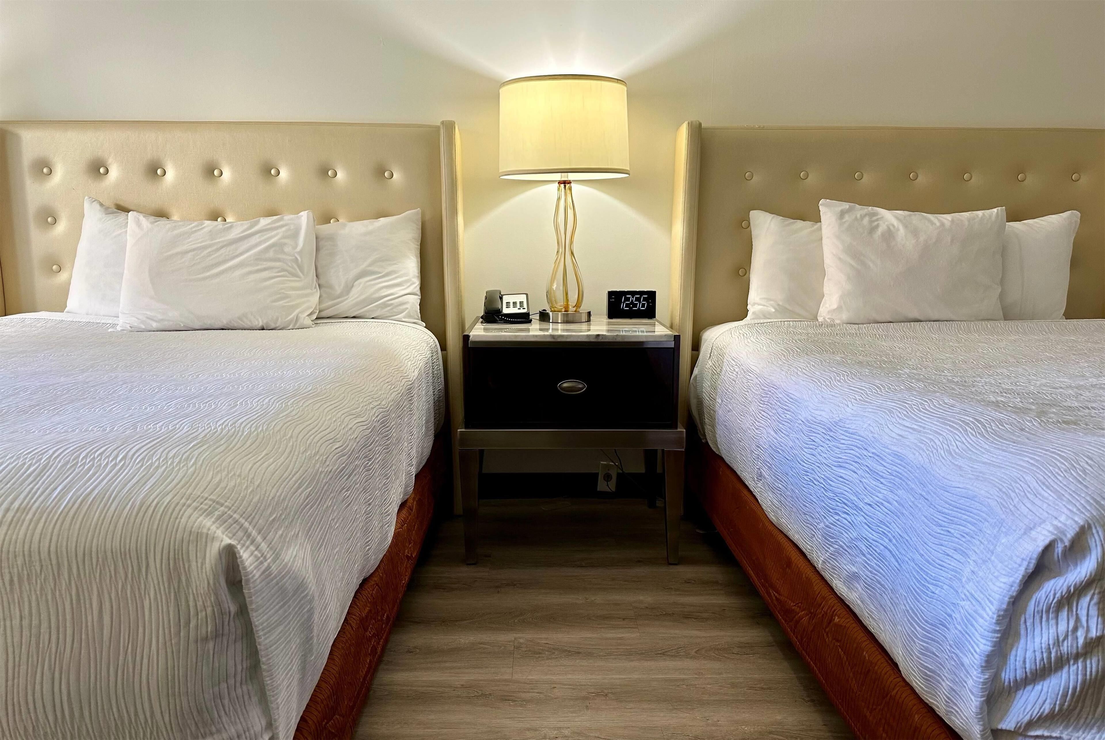 A hotel room with two beds, a nightstand with a lamp, a clock, and a phone.