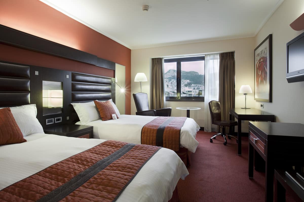 A hotel room with two beds, a desk, a chair, a TV, and a window with a city view.