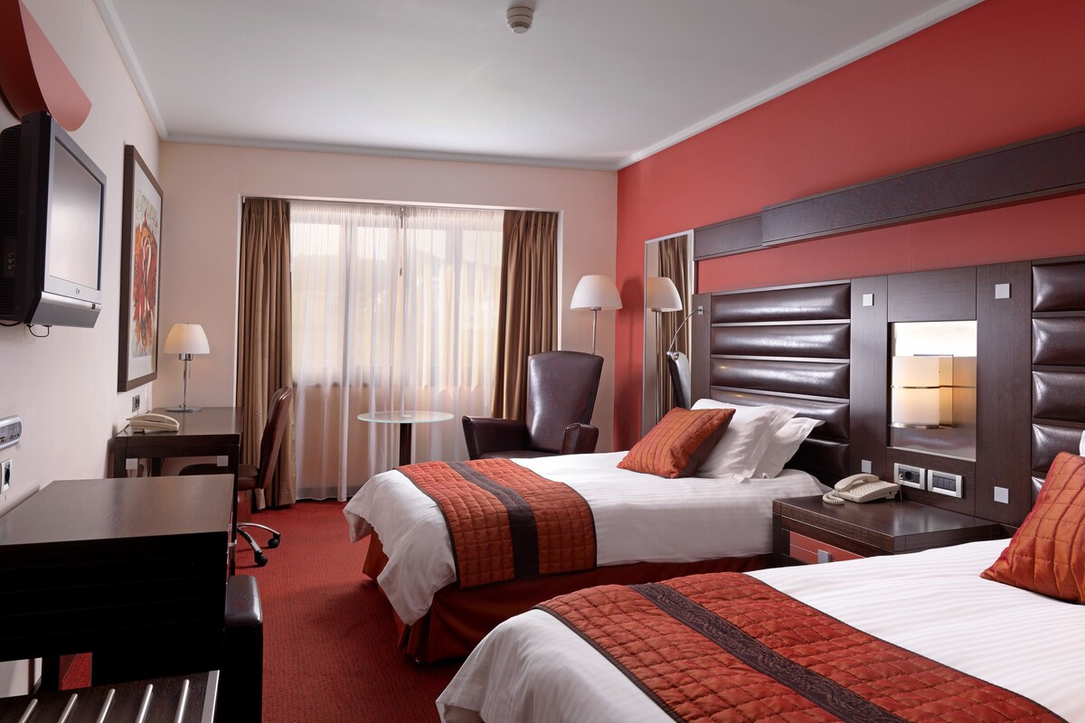 A hotel room with two beds, a desk, a chair, a TV, and a window with curtains.