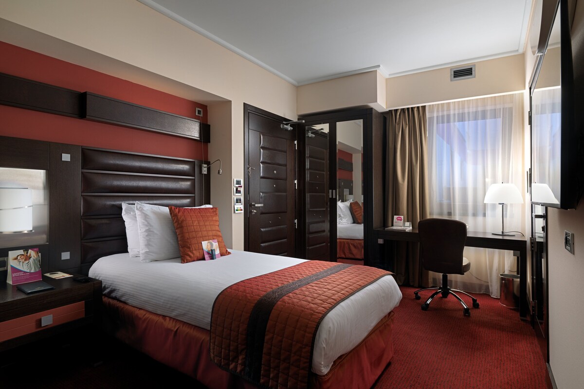 A hotel room with a large bed, a desk, a chair, a TV, and a mirror.