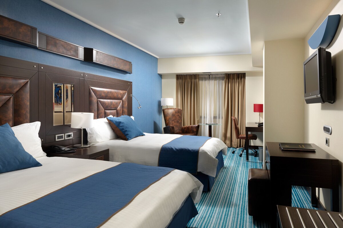 A hotel room with two beds, a desk, a chair, a television, and a window with curtains.