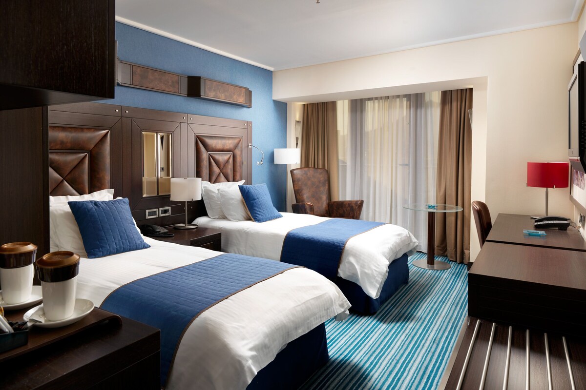 A hotel room with two beds, a desk, a chair, a TV, and a window with curtains.
