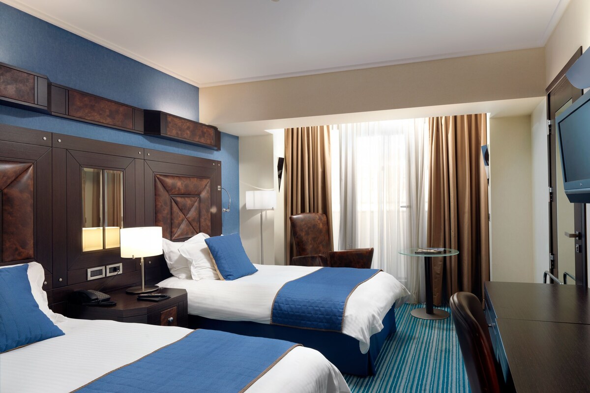 A hotel room with two beds, a desk, a chair, a TV, and a window with curtains.