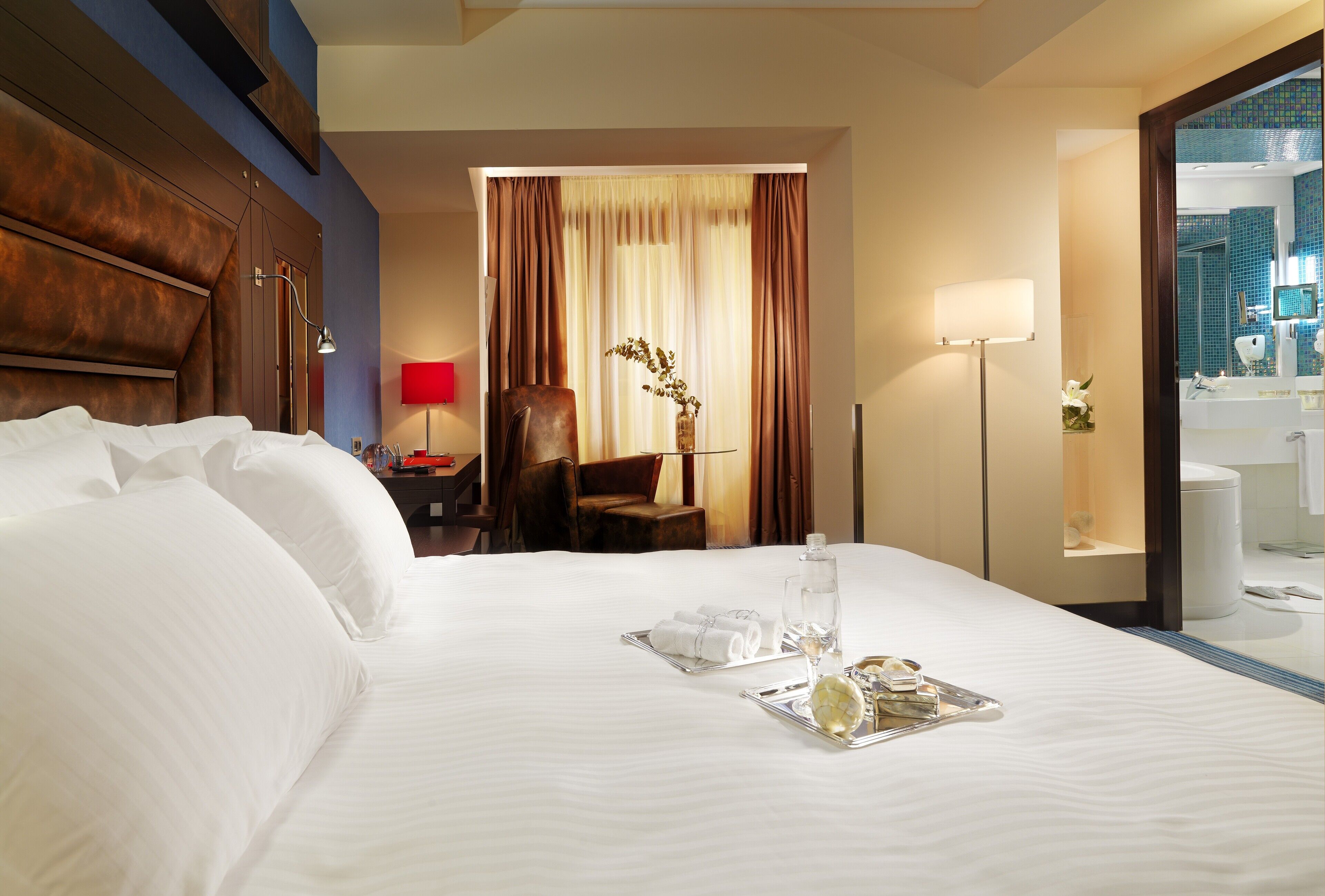 A hotel room with a large bed, a desk with a chair, a lamp, and a bathroom visible through a glass door.
