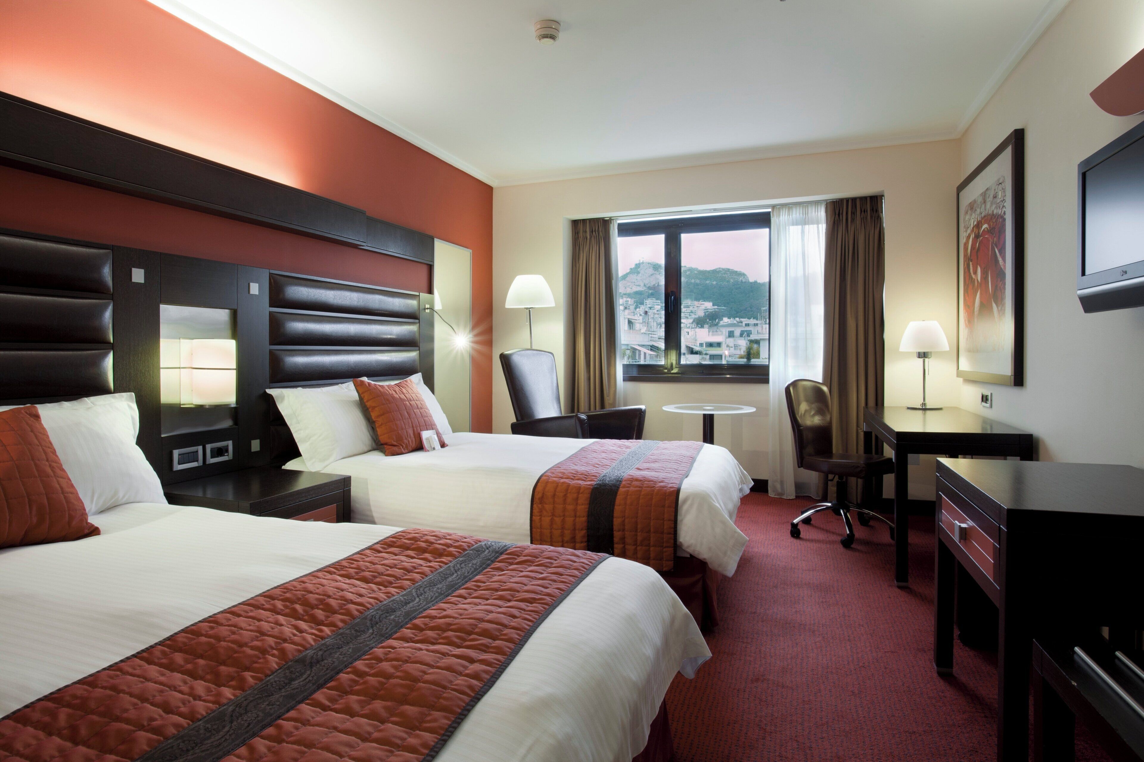 A hotel room with two beds, a desk, a chair, and a window with a city view.