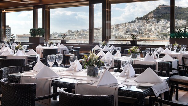 Un restaurant avec des tables dressées pour le service, de grandes fenêtres donnant sur la ville et des chaises en osier.