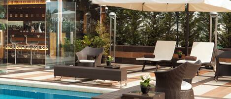 Un área junto a la piscina con sillones de descanso, una mesa y una sombrilla grande.