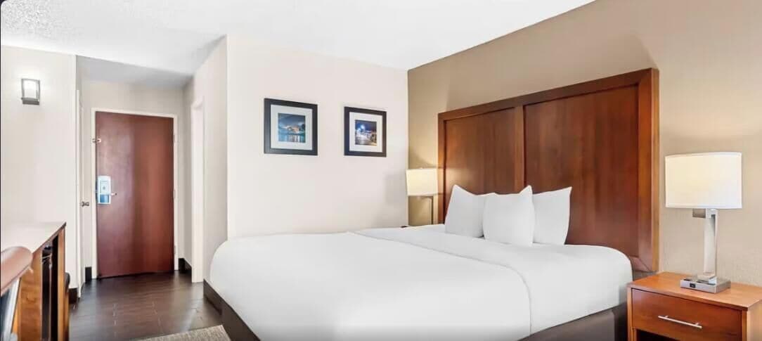 A hotel room with a large bed, a wooden headboard, two bedside lamps, and a door leading to another room.