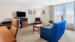 A hotel room with a sofa, a wooden table, a TV, a desk with drawers, a lamp, and a window with curtains.