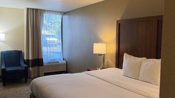 A hotel room with a large bed, a chair, a desk with a lamp, and a window with curtains.