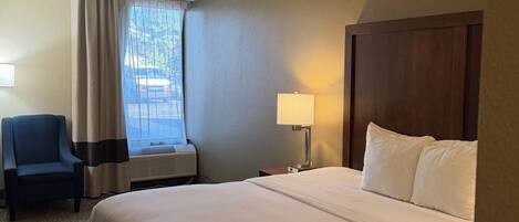 Una habitación de hotel con una cama grande, una silla, un escritorio con lámpara y una ventana con cortinas.