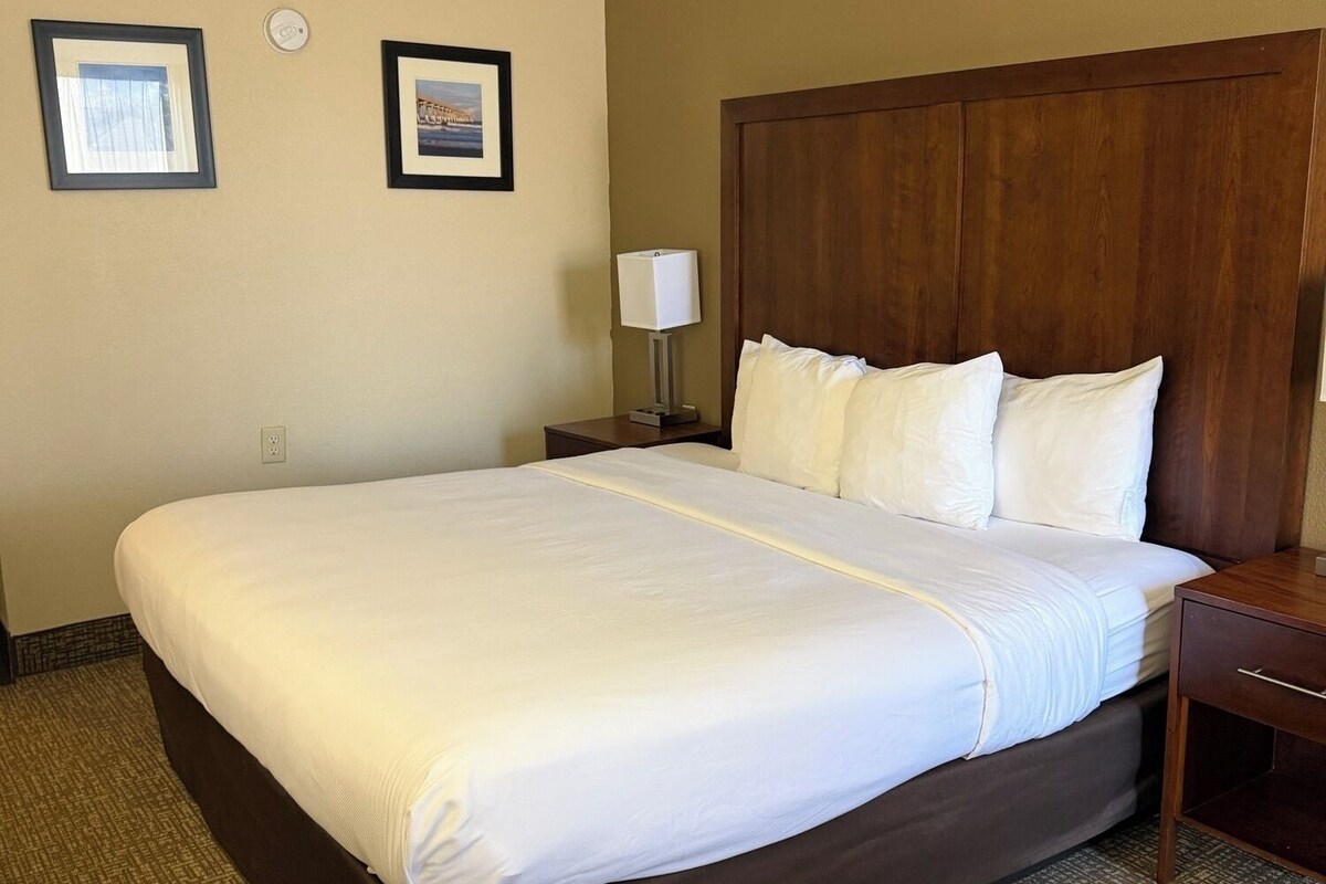 A hotel room with a large bed, a wooden headboard, two framed pictures on the wall, a bedside table with a lamp, and a carpeted floor.