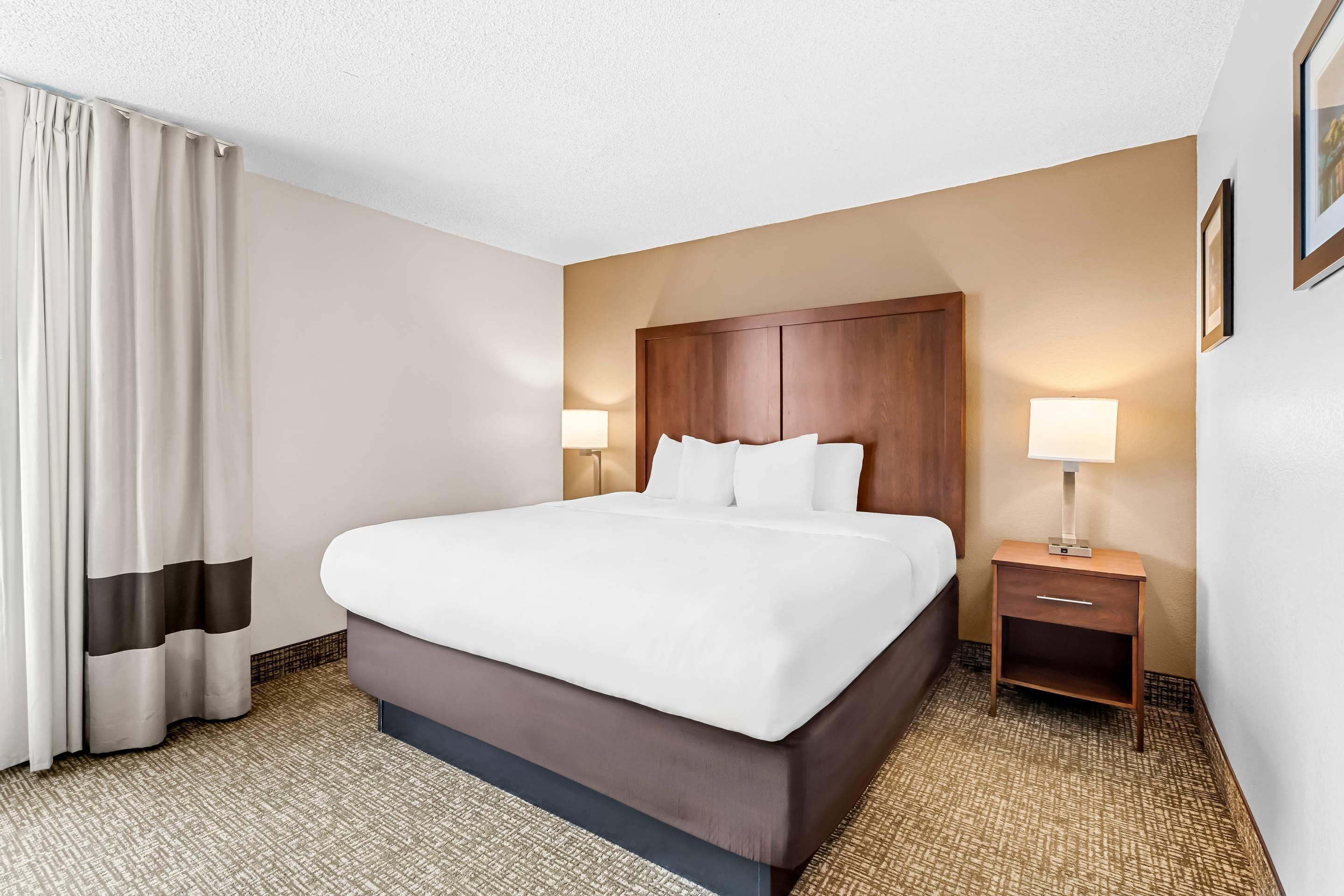 Una habitación de hotel con una cama grande, una mesita de noche, una lámpara y una ventana con cortinas.