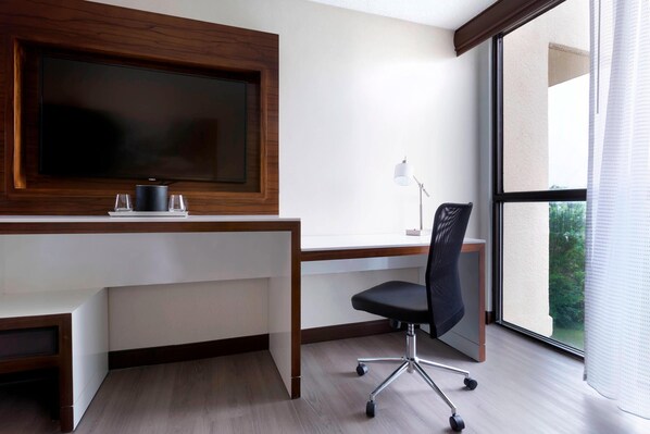 A modern office space with a large flat-screen TV mounted on a wooden wall, a white desk, a black office chair, and a large window offering a view of greenery. - Marriott Greensboro Airport (Greensboro)