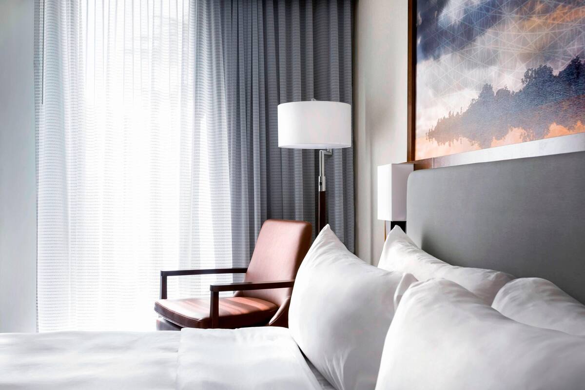 A hotel room with a bed, a chair, a lamp, and a painting on the wall.