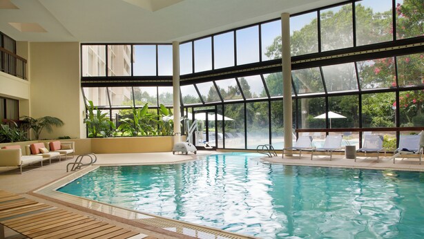 An indoor swimming pool with clear glass walls, lounge chairs, and a view of the outdoors.