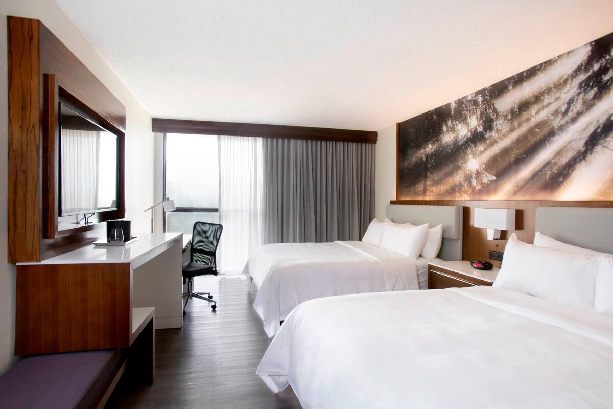 A hotel room with two beds, a desk, a chair, a large window with curtains, and a painting on the wall.