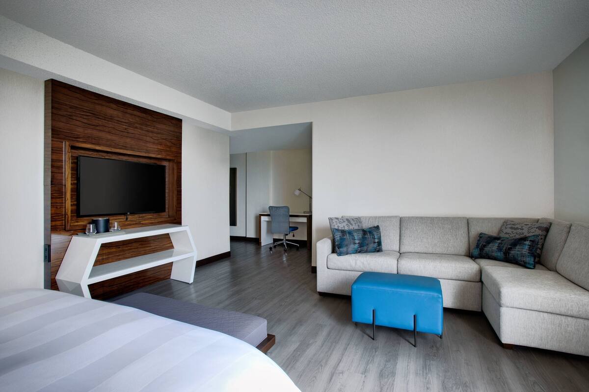A modern hotel room with a large bed, a sofa, a TV mounted on a wooden panel, and a desk with a chair.