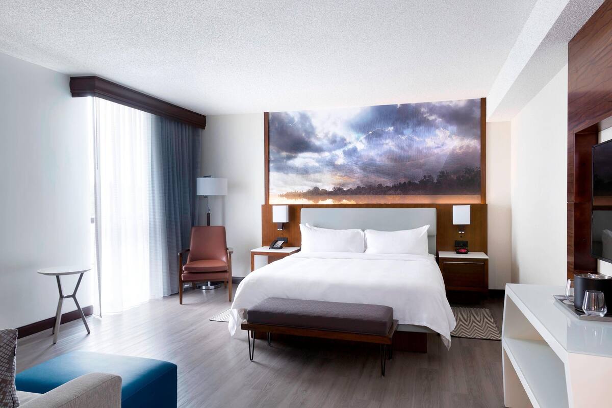 A hotel room with a large bed, a desk, a chair, a bench, and a large painting of a landscape.