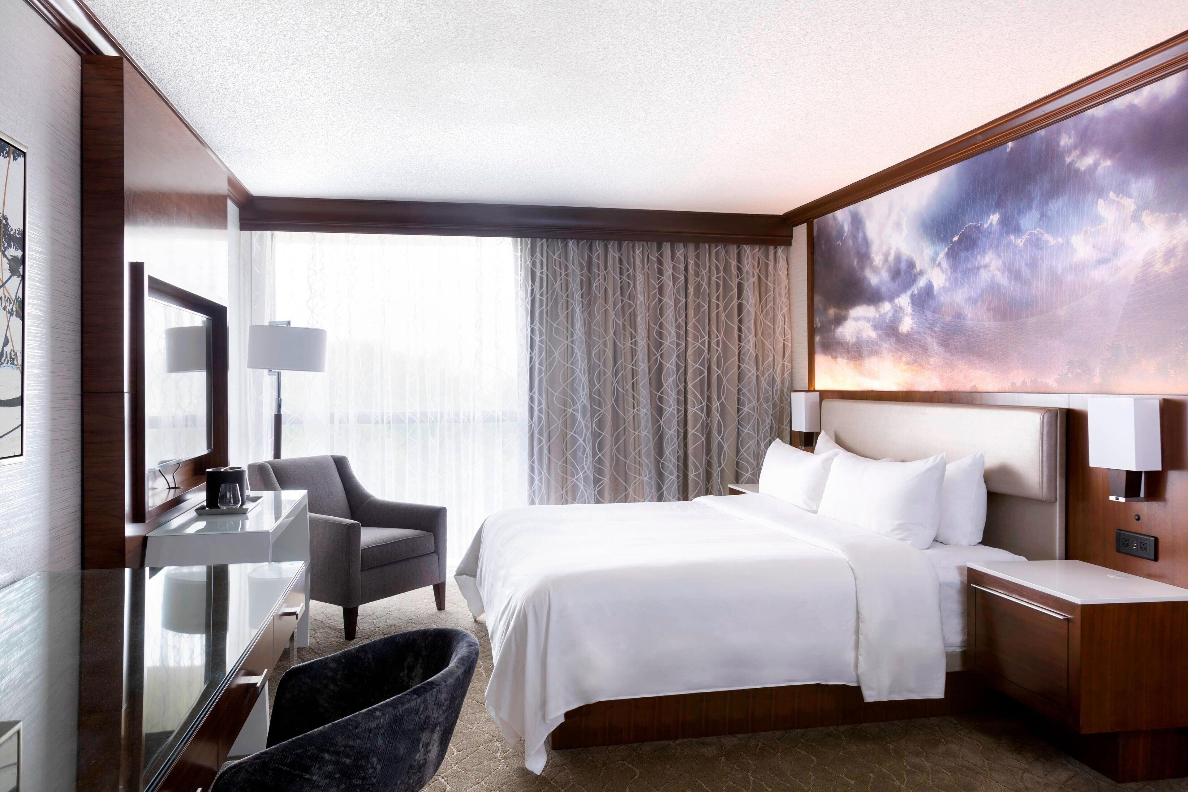 Une chambre d’hôtel avec un grand lit, un bureau, une chaise, une table de chevet et une grande fenêtre avec des rideaux.