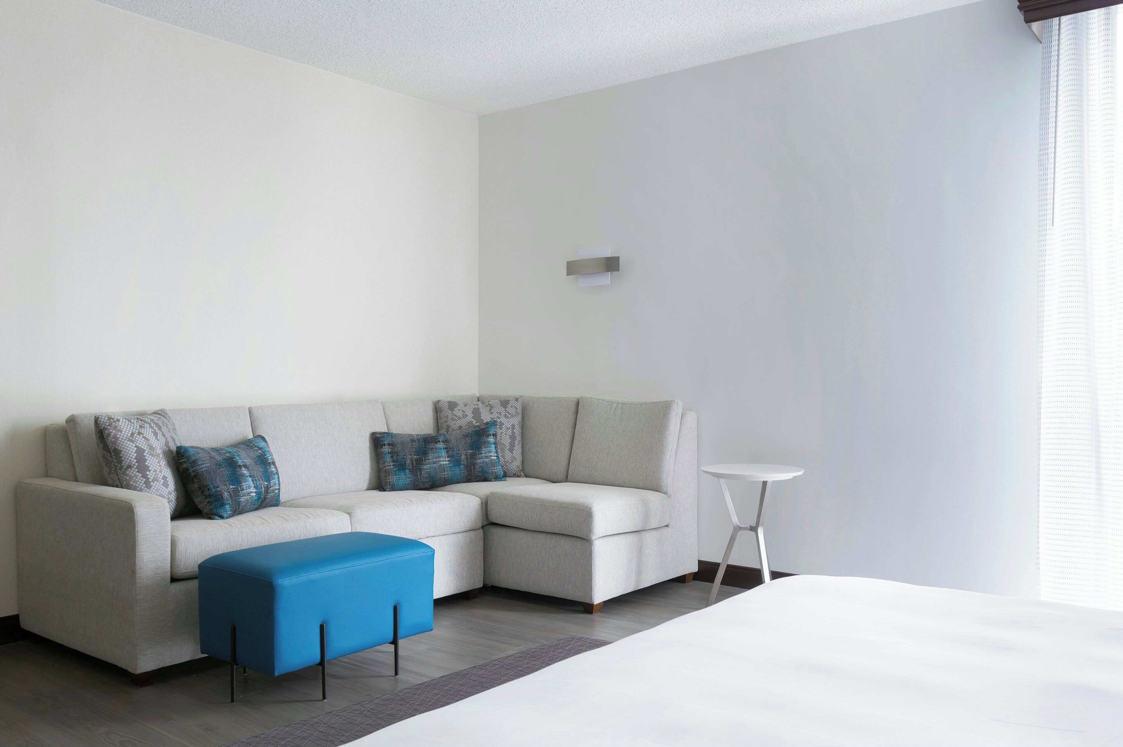 Un salon moderne avec un canapé d’angle, un pouf bleu, une petite table d’appoint blanche et une grande fenêtre équipée de stores.