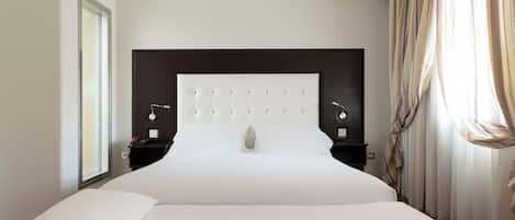 Una habitación de hotel moderna con una cama grande, cabecera oscura, mesitas de noche con lámparas y un amplio ventanal con cortinas.