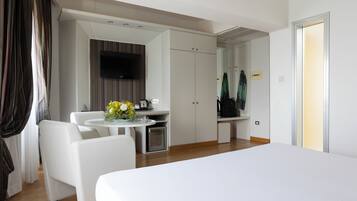 A modern hotel room with a bed, a dining table with chairs, a small kitchenette, and a TV mounted on the wall.
