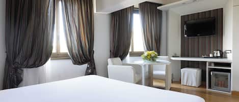 Habitación de hotel con una cama grande, una mesa pequeña con sillas, un televisor montado en la pared y un minibar.