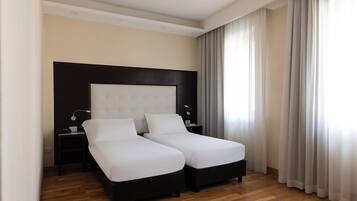 A modern hotel room with two single beds, wooden flooring, a large headboard, bedside tables with lamps, and sheer curtains.