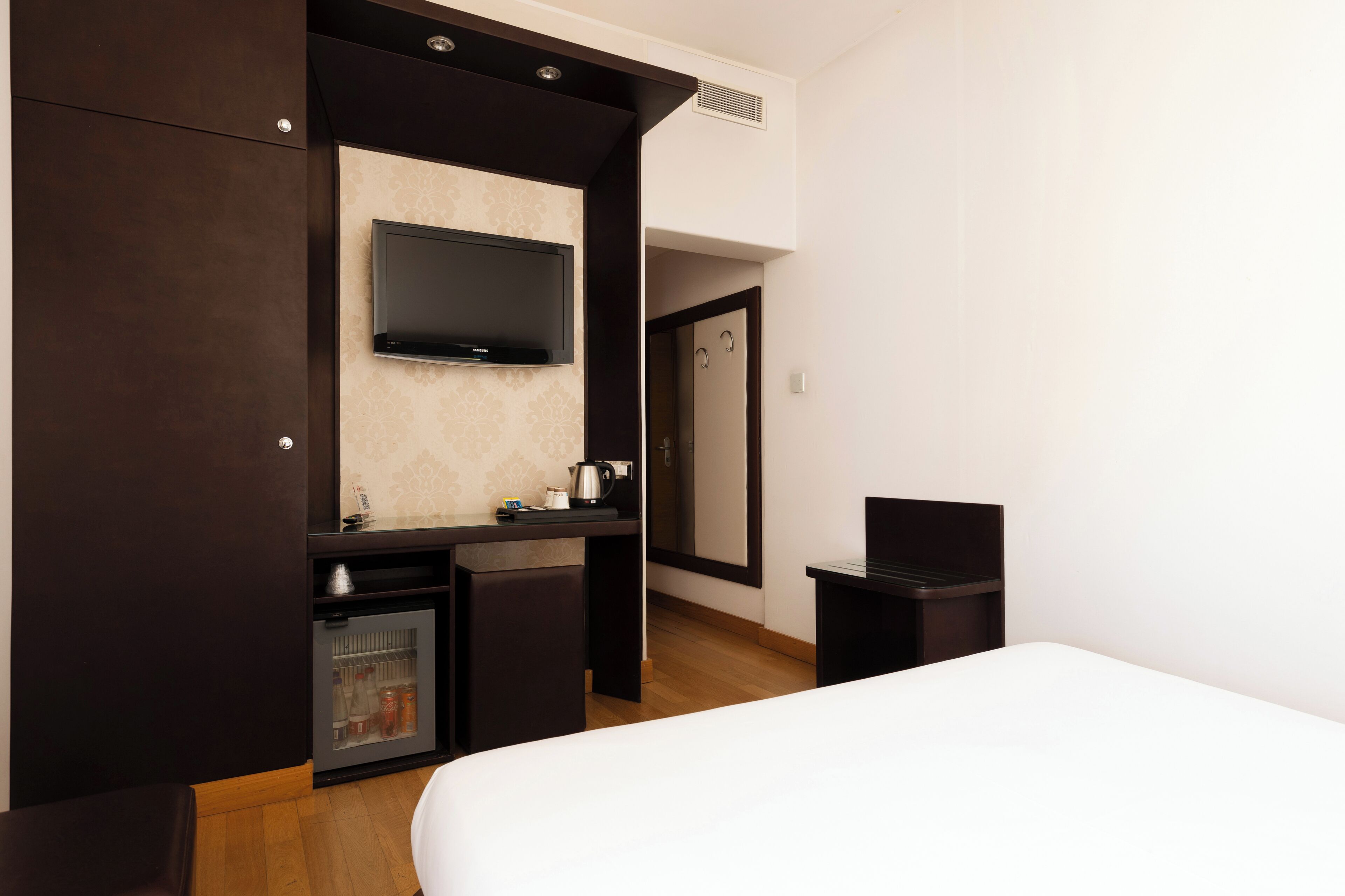 A hotel room with a bed, a TV mounted on the wall, a small kitchenette with a fridge, and a door leading to the bathroom.