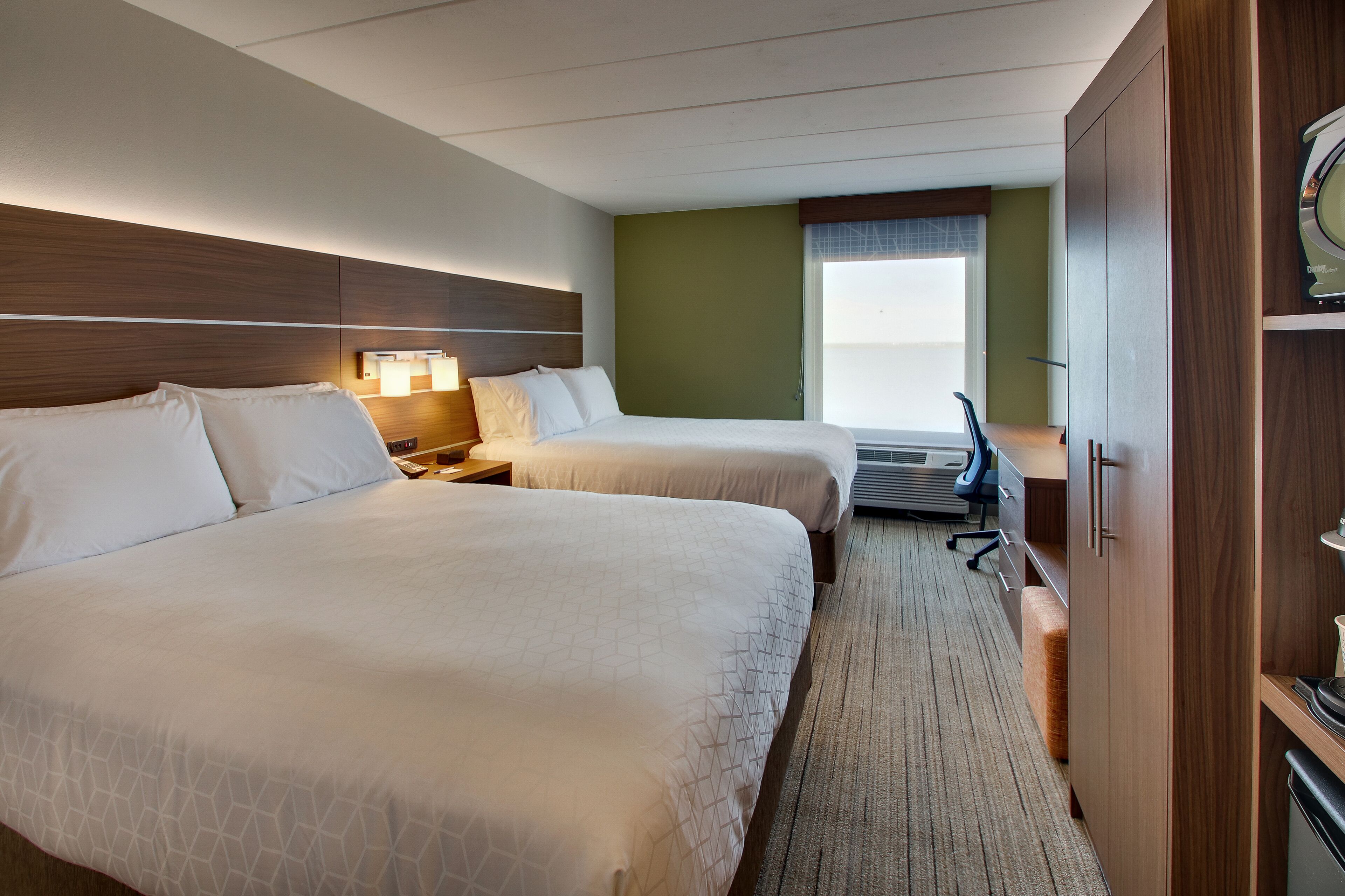 Une chambre d’hôtel avec deux lits, un bureau, une chaise, une lampe et une fenêtre avec des stores.