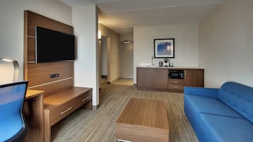 A hotel room with a flat-screen TV, a sofa, a coffee table, a desk, and a kitchenette.