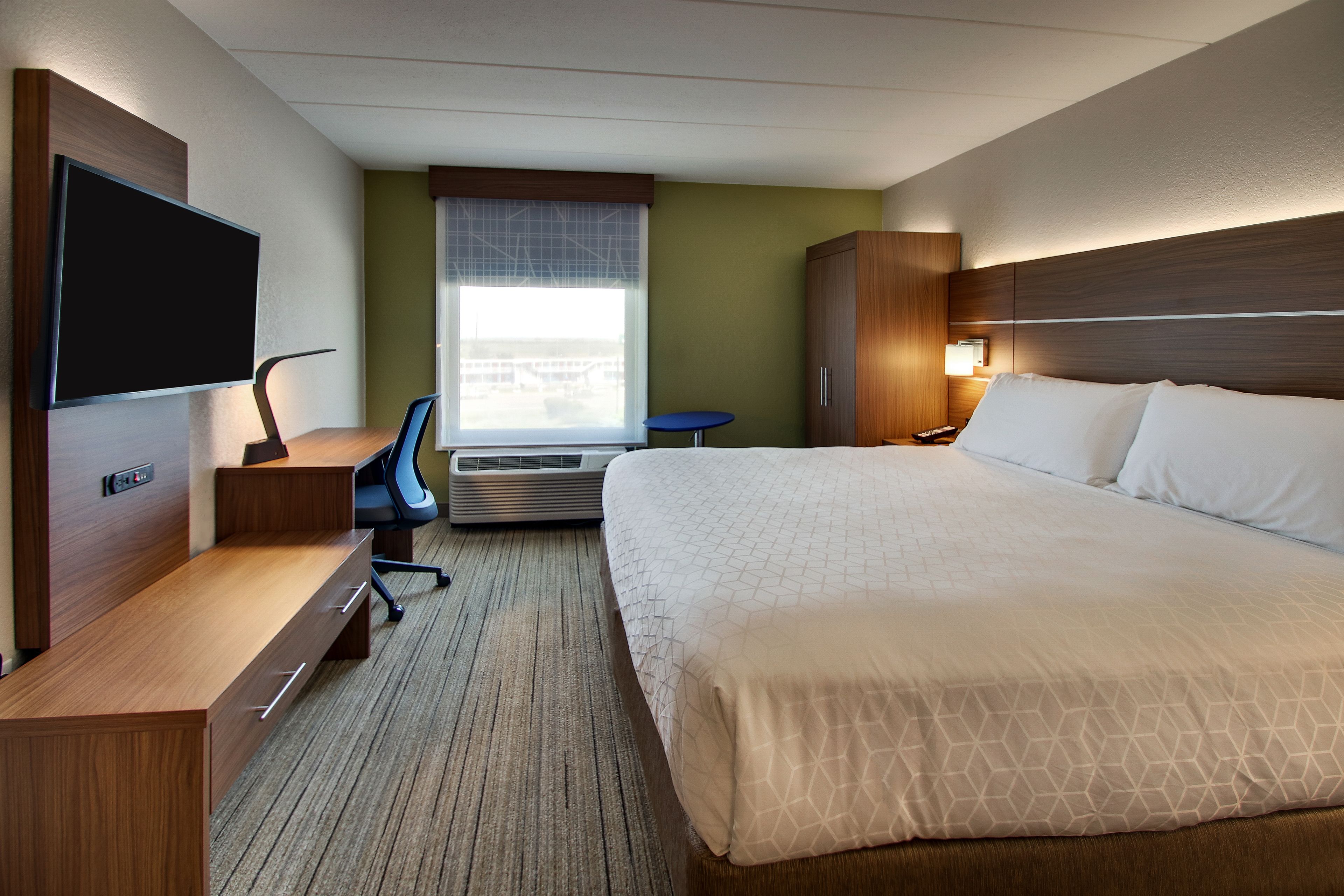 A hotel room with a bed, a desk with a chair, a television, an air conditioning unit, and a window with blinds.