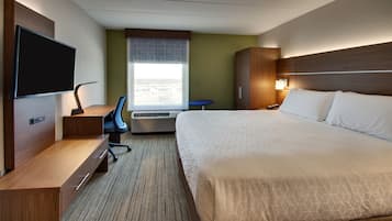 A hotel room with a bed, a desk with a chair, a television, an air conditioning unit, and a window with blinds.