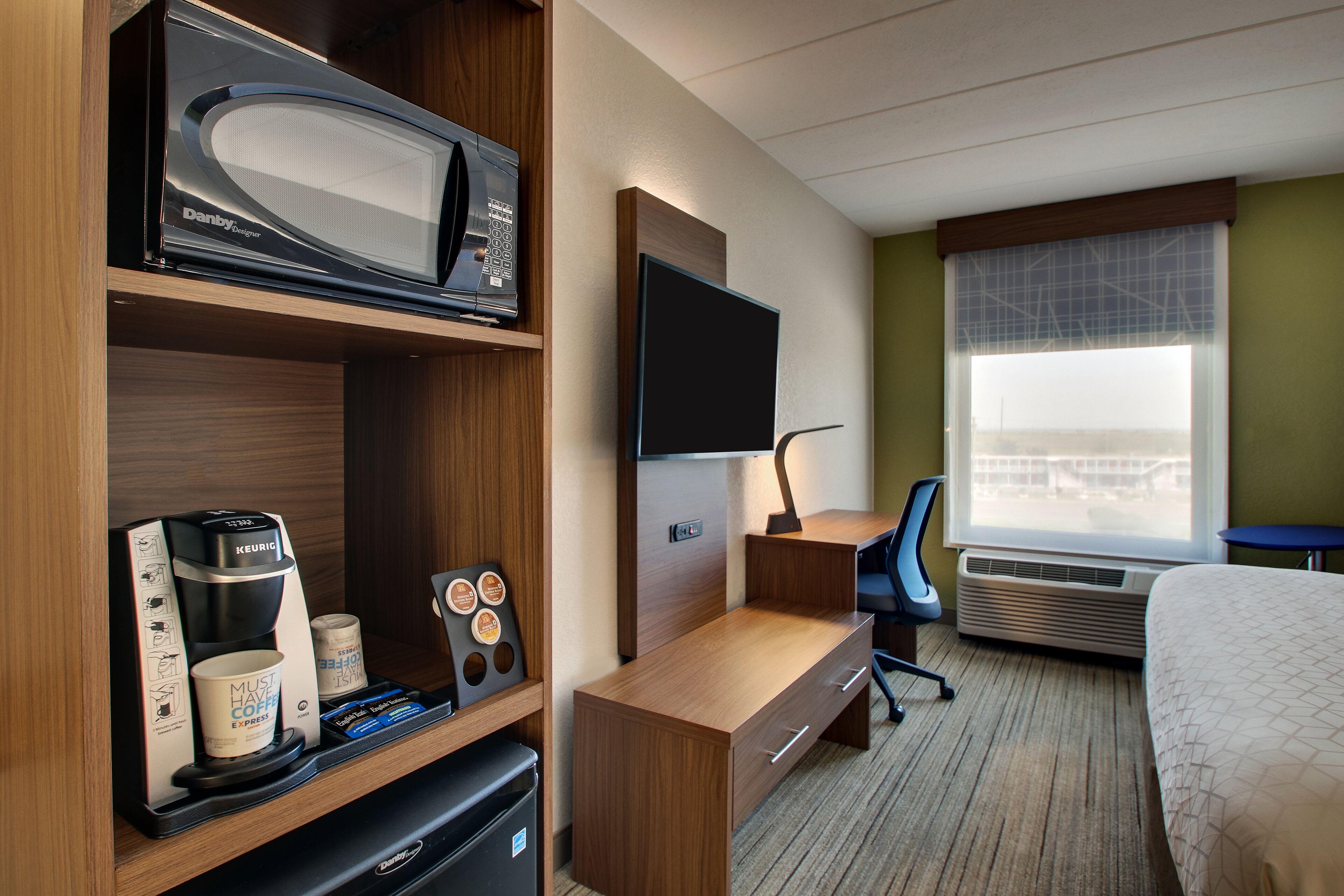 A hotel room with a microwave, coffee maker, TV, desk, chair, and a bed.