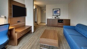 A modern hotel room with a flat-screen TV mounted on a wooden panel, a blue sofa, a wooden coffee table, a desk with a chair, and a kitchenette area with a microwave and coffee maker.